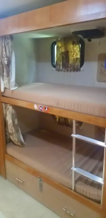 This RV rental features two bunks with light brown mattresses. Wooden frames, patterned curtains, a window, and a ladder are visible. Storage drawers are below.. Winnebago Sightseer 2007