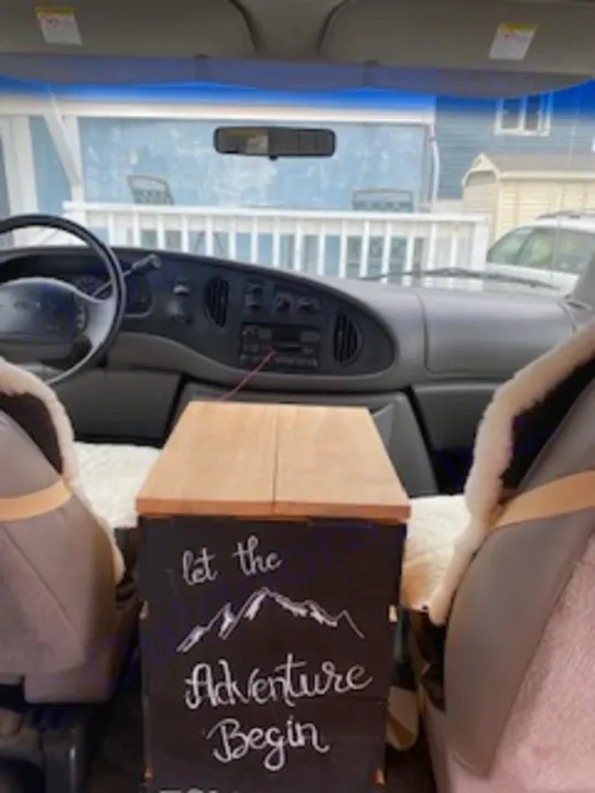 Interior view of an RV rental van, looking forward. Features front seats, dashboard, and a decorative "Let the Adventure Begin" sign.. Ford Econoline E250supercab 1999