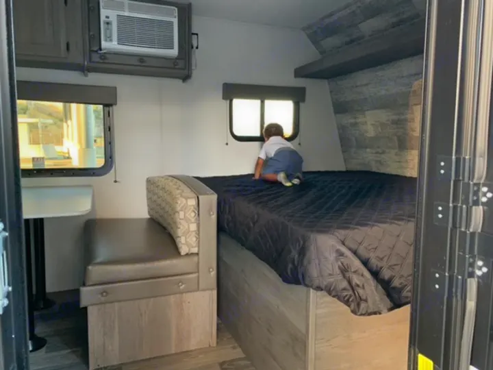 Inside a compact RV rental, a child explores the bed next to a dinette. An AC unit is mounted above.. Keystone Springdale 2021