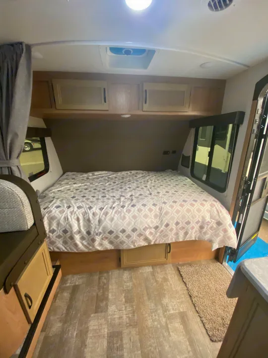Interior of an RV rental: a comfortable bed with a patterned comforter, overhead cabinets, and windows.. Winnebago Micro Minnie 2021
