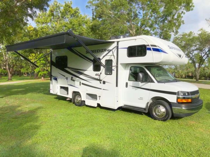 White motorhome RV rental with its awning extended, parked on green grass in a sunny park setting. Ready for your adventure!. Forest River sunseeker LE 2350 2020