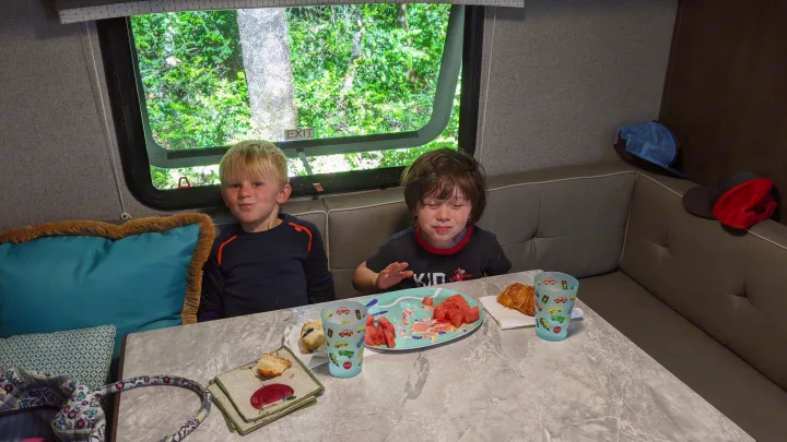 Two young boys have breakfast inside an RV rental, enjoying watermelon and pastries by a leafy window.. Forest River Rockwood Mini Lite 2020