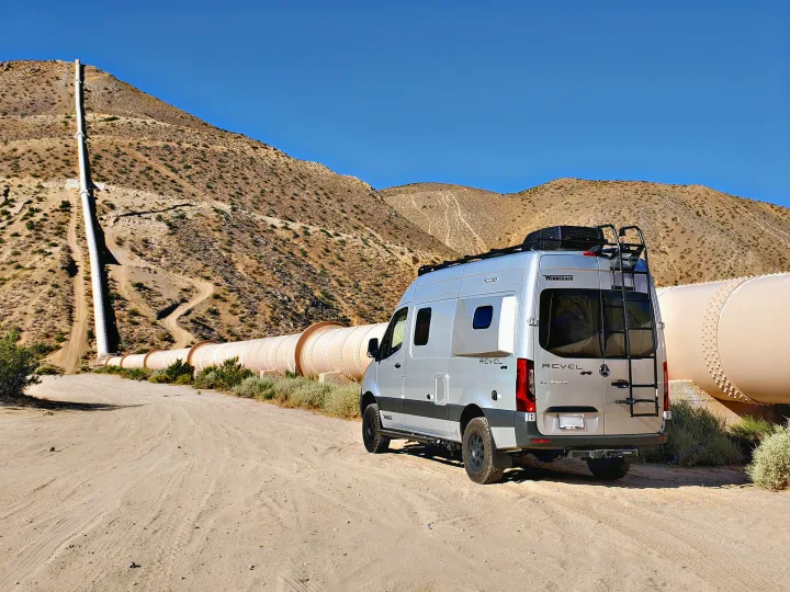 Explore the desert! This silver Winnebago Revel 4x4 camper van, ideal as an rv rental, rests by a vast pipeline and mountains.. Winnebago Revel 2020