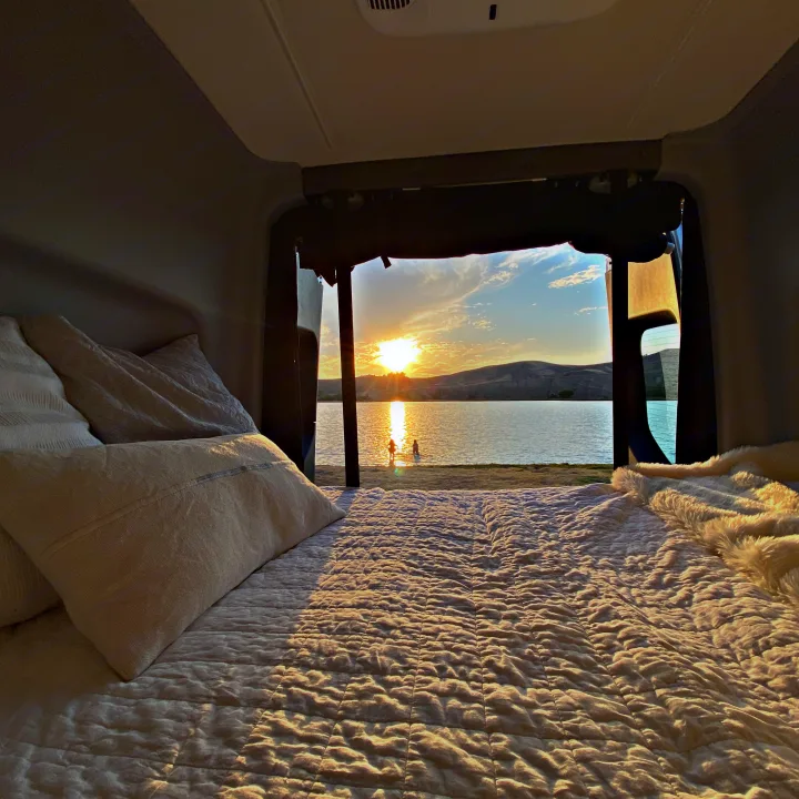 Inside your cozy RV rental, enjoy a sunset view of a lake with two people wading. A comfy bed awaits.. Winnebago Revel 2020