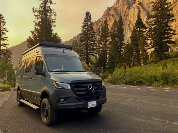 Grey Mercedes Sprinter RV rental on a mountain road, surrounded by tall pines and rocky peaks. Ideal for adventure.. Winnebago Revel 2021
