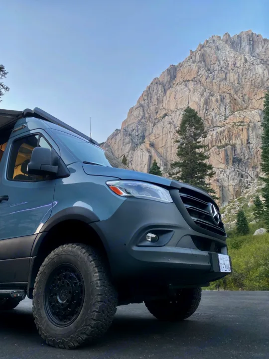 Adventure-ready Mercedes Sprinter van on pavement, with a massive rocky mountain & trees in the background. Ideal rv rental.. Winnebago Revel 2021
