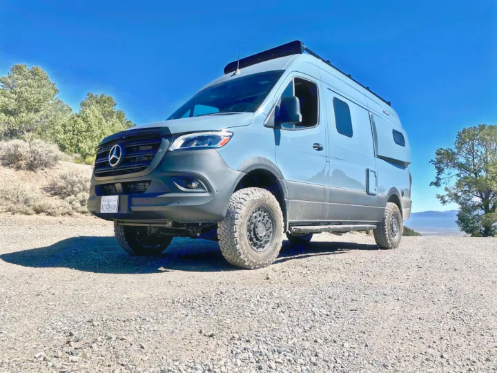 A light blue Mercedes Sprinter adventure van, an ideal rv rental, sits on a gravel road with distant mountains and a clear sky.. Winnebago Revel 2021