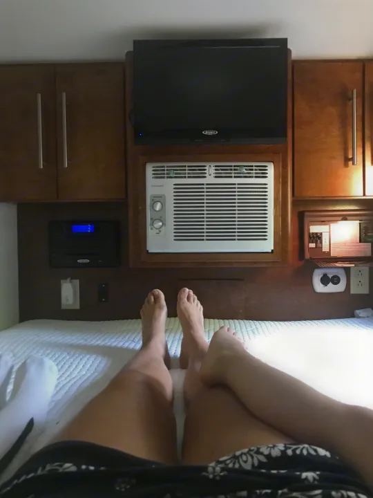 Relaxing in an rv rental bedroom: POV from the bed, showcasing legs, feet, and a wall unit with TV, AC, and control panel.. T@G T@G Trailer 2015