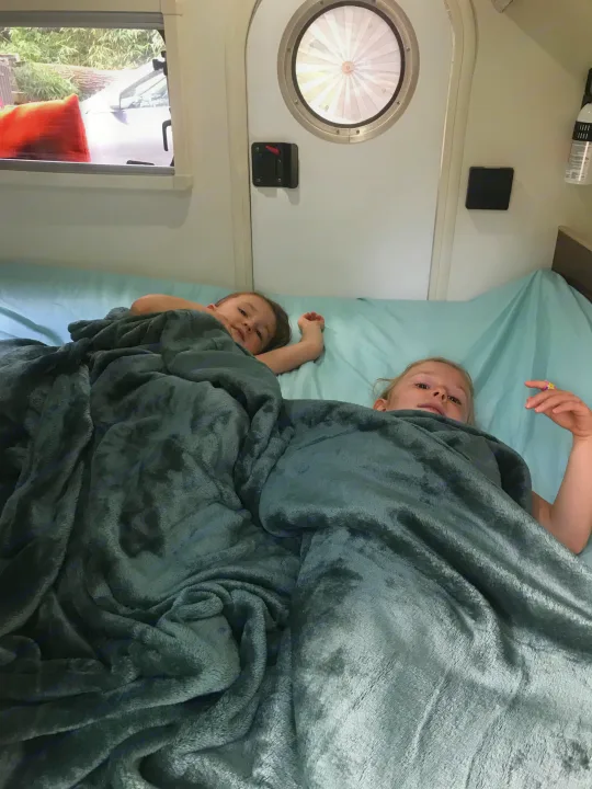 Two children cozy in their RV rental bed. They lie under soft gray blankets in the camper, looking at the camera.. T@G T@G Trailer 2015