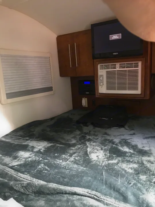 RV rental interior: cozy bedroom with a grey blanket on the bed, window, TV, AC unit, and wooden storage cabinets.. T@G T@G Trailer 2015