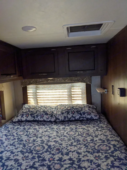 Comfortable rv rental bedroom with floral bedding, dark cabinets, a sunlit window, and ceiling vent.. Other Other 2020