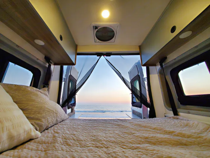 Interior of an RV rental: a plush bed faces open back doors, offering a serene beach and ocean sunset vista.. Winnebago Solis 2021
