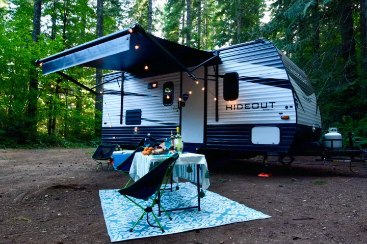 This RV rental campsite features a white and blue travel trailer with an extended awning, string lights, and outdoor dining.. Keystone Hideout 2021