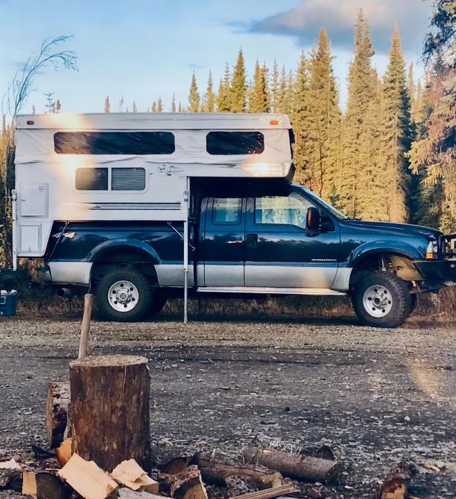 Blue truck with Bronco pop-up truck camper, an RV rental, at a forest campsite with firewood. Perfect for outdoor adventures.. Ford F250 2005