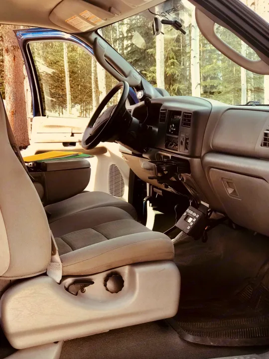 Inside a roomy RV rental: driver's seat, dashboard with radio, and cup holder visible. Trees outside for your travel adventure.. Ford F250 2005