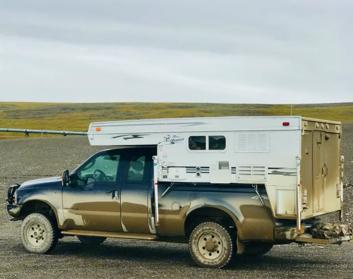 A robust, mud-splattered pickup truck with a mounted Bronco truck camper. Ideal for an adventurous RV rental exploring remote landscapes.. Ford F250 2005