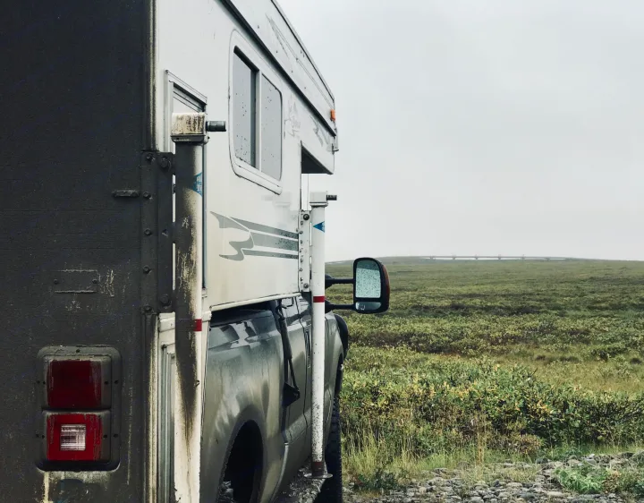 Mud-splattered RV rental parked in vast green tundra under a cloudy sky, with a distant pipeline.. Ford F250 2005