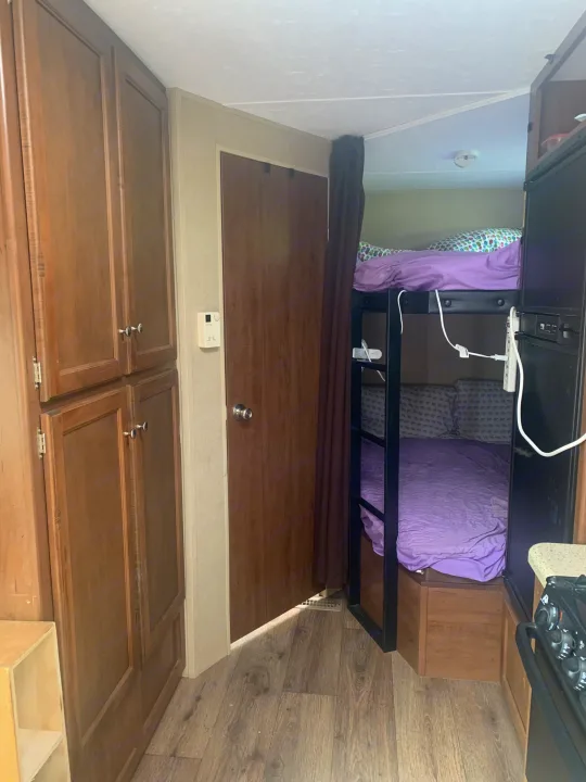 Compact RV rental interior shows wooden storage cabinets, a door, and bunk beds with purple bedding near a small kitchen.. Dutchmen Aspen Trail 2014
