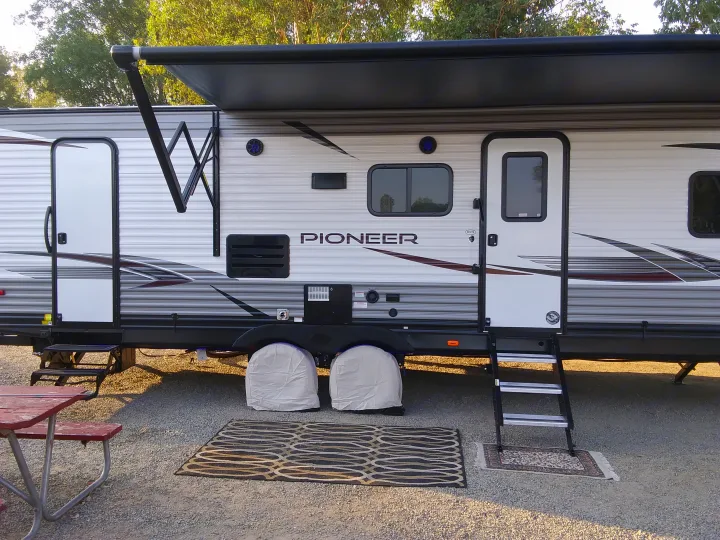 Side view of an RV rental travel trailer with its black awning extended, steps down, an outdoor rug, and a picnic table.. Heartland Pioneer 2021