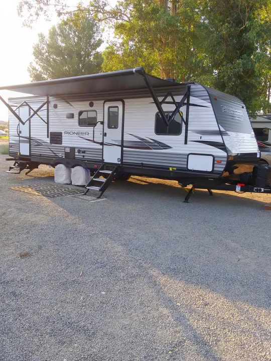 A "PIONEER" RV rental travel trailer parked on a gravel site with its awning extended.. Heartland Pioneer 2021