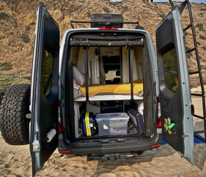 Open doors of a cool rv rental camper van on sand, showing a cozy bed & gear. Perfect for your next road trip adventure!. Winnebago Revel 2021