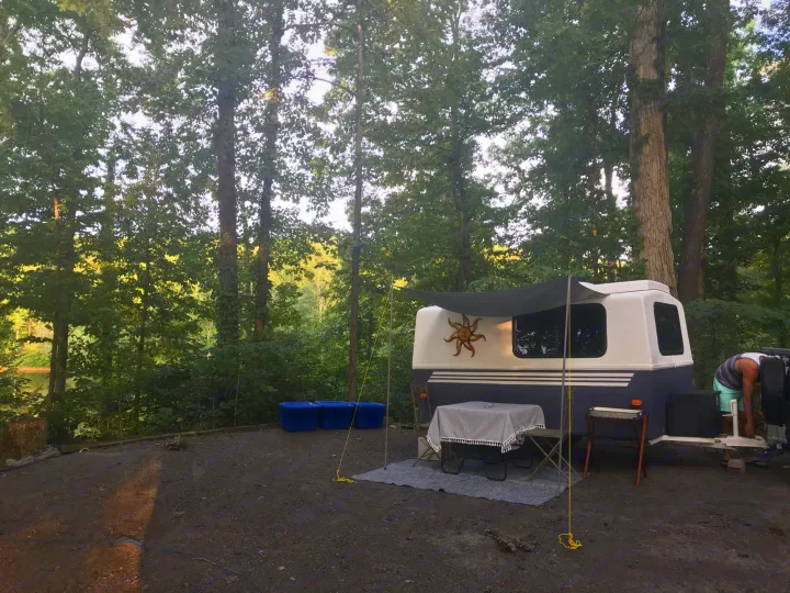 Enjoying an rv rental at a scenic wooded campsite. White/blue trailer, awning, table, and person setting up by a lake.. Hunter Compact Junior 1971