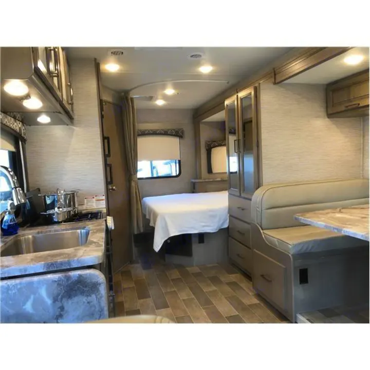 Interior view of a modern RV rental. It shows a kitchen with a sink, a bed with white linen, and a beige dining booth.. Thor Motor Coach Chateau 2020