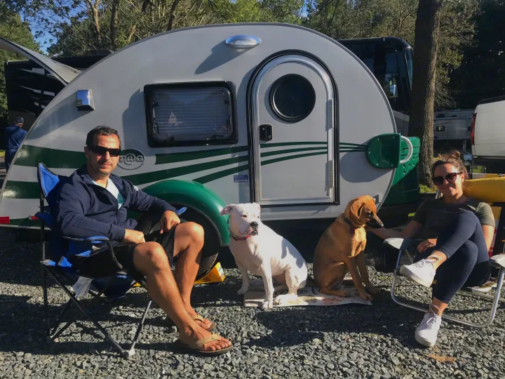 A couple and two dogs relax outside their teardrop RV rental at a sunny campsite, enjoying outdoor leisure.. T@G T@G Trailer 2015