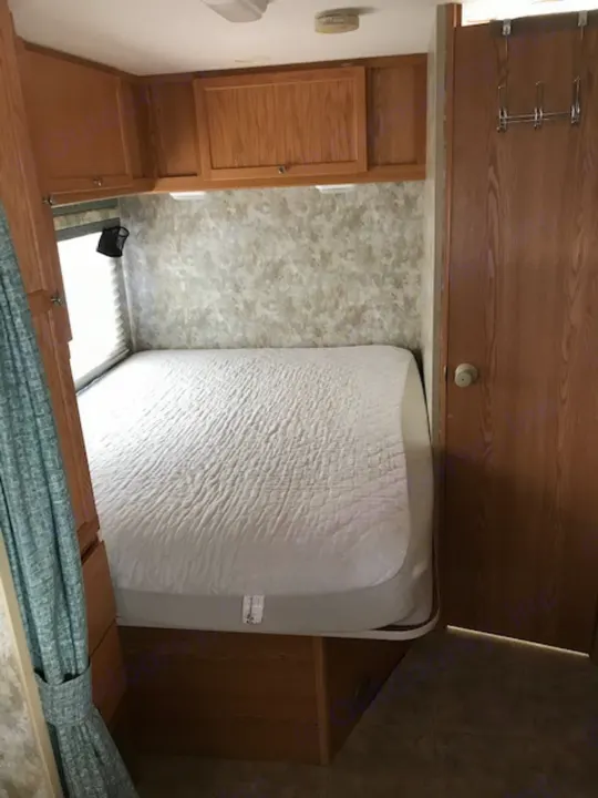 Compact RV rental bedroom featuring a comfortable bed with white bedding, integrated wood cabinets, and patterned flooring.. Winnebago Access 2007