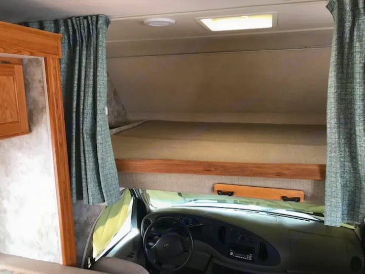 View inside an RV rental showing the elevated over-cab bunk bed, with the vehicle's dashboard and steering wheel below.. Winnebago Access 2007