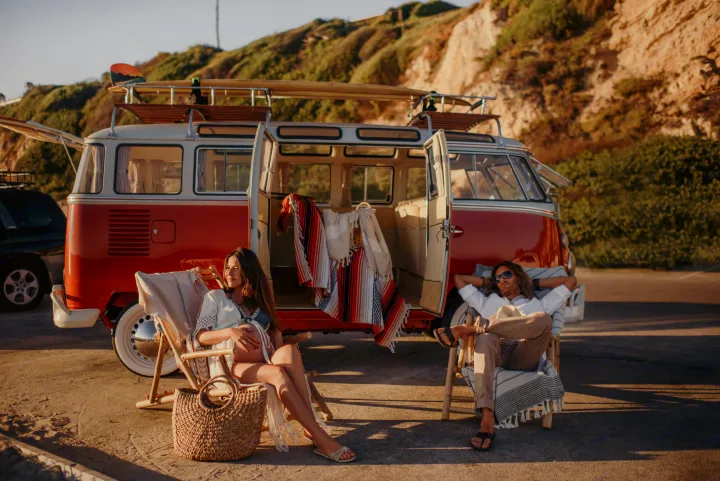 A couple relaxes beside their vintage red VW camper van with surfboards. Perfect for your next RV rental adventure!. Volkswagen Kombi 1965
