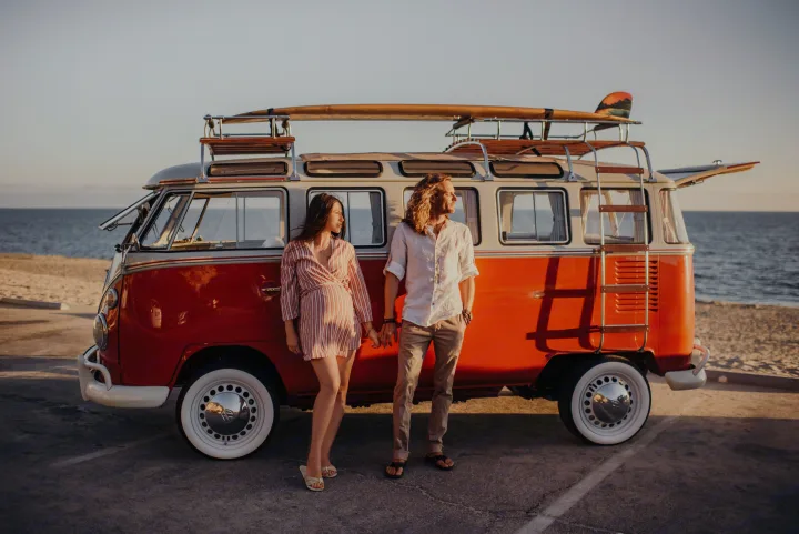 A couple by a vintage VW camper van with a surfboard, parked at the beach. Ready for your next RV rental adventure!. Volkswagen Kombi 1965