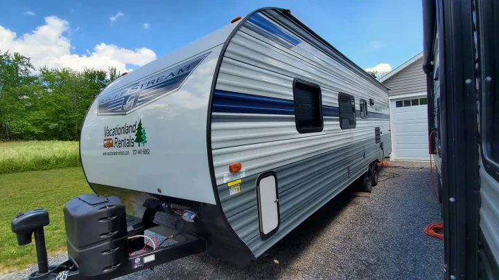 Side view of a Gulf Stream travel trailer RV rental, white & gray, with "Vacationland Rentals" logo, parked on gravel.. Gulf Stream Trailmaster 2021