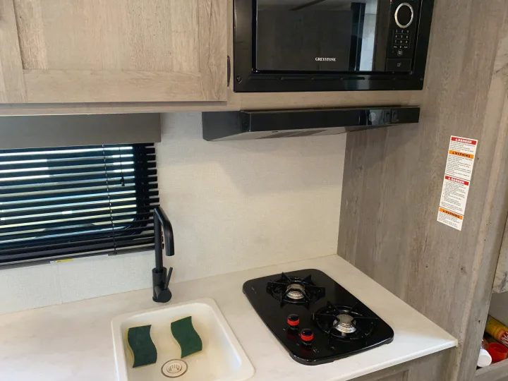 This rv rental kitchen features a Greystone microwave, two-burner cooktop, and sink with sponges. A compact camper galley.. Keystone Springdale 2021