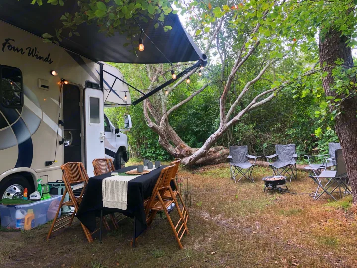 An RV rental campsite setup, featuring an extended awning with string lights, outdoor dining, and a cozy fire pit area.. Other Four Winds 2018