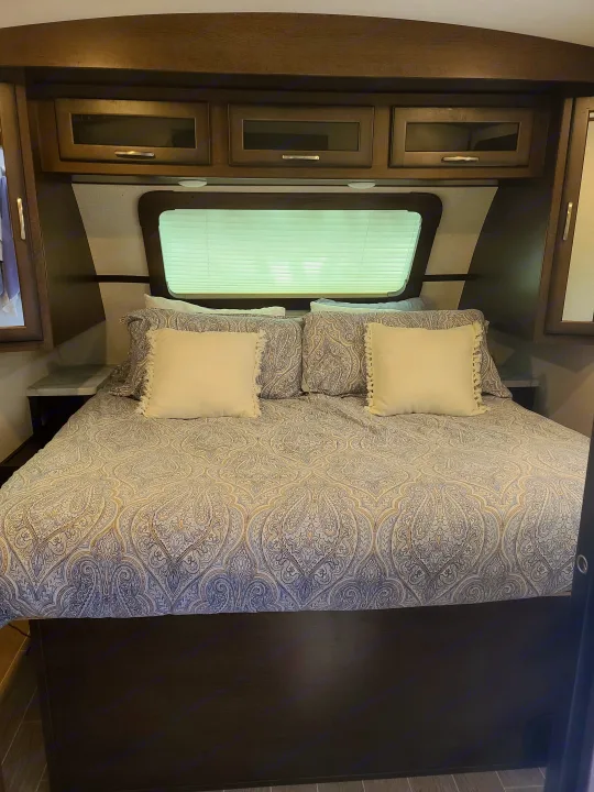 Comfortable RV rental bedroom featuring a bed with patterned paisley bedding, white pillows, and built-in wood cabinets and window.. Dutchmen Other 2020