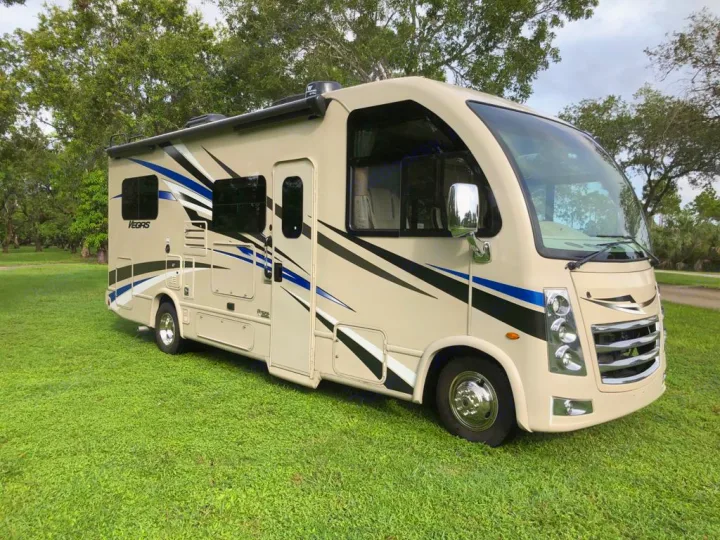 This cream-colored Class C RV rental, featuring black and blue graphics, is parked on green grass. Ideal for your next road trip.. Thor Motor Coach Vegas 2020
