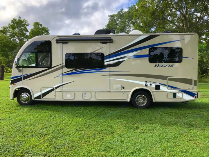 Side view of a beige Thor Vegas RV rental, a spacious motorhome parked on green grass under a cloudy sky.. Thor Motor Coach Vegas 2020