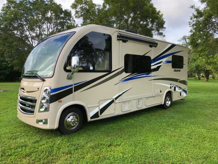 This beige Class A RV rental, adorned with blue & black graphics, is parked on lush green grass, ready for your next adventure.. Thor Motor Coach Vegas 2020
