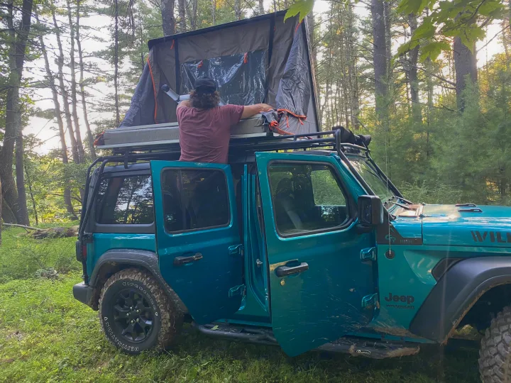 A person readies a rooftop tent on a teal Jeep Wrangler, offering a unique camping rv rental alternative in the woods.. Other Other 2020