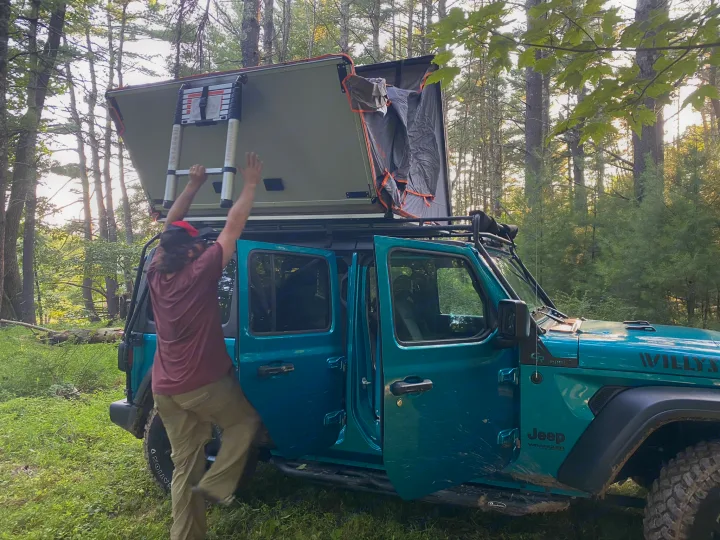 Person deploying a rooftop tent on a teal Jeep Wrangler, ideal for a rugged RV rental camping experience.. Other Other 2020