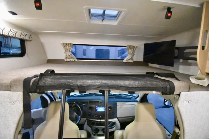An RV rental's interior: a cab-over bunk with safety net, mounted TV, and the vehicle's dashboard.. Leprechaun 230RB 2020