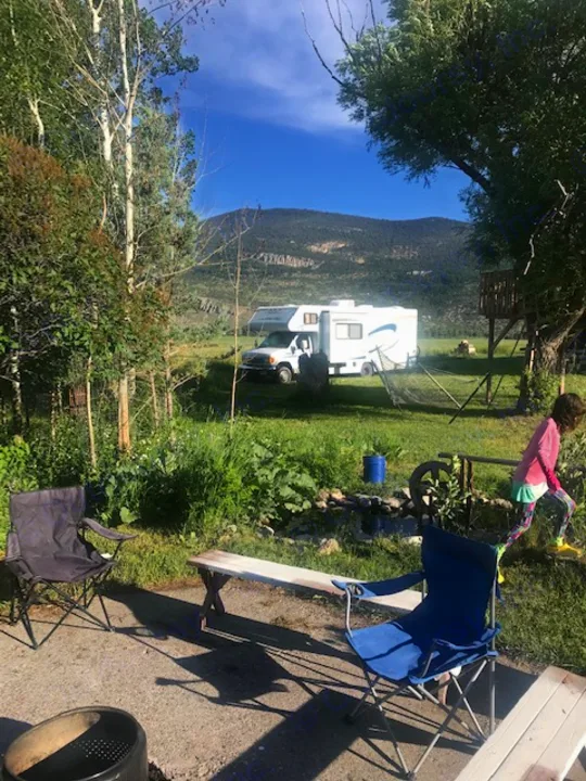 An RV rental is set up at a scenic mountain campsite with a child, chairs, and a bench. Ideal for outdoor adventures.. Winnebago Access 2007