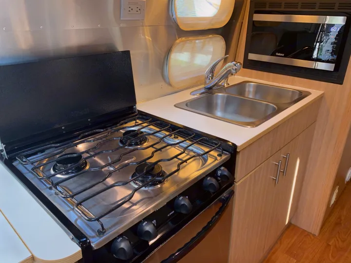 This RV rental kitchenette features a stainless steel stove/oven, double sink, and microwave, with light wood cabinets and metallic walls.. Airstream Safari 2006