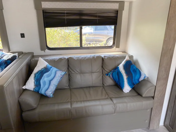 Inside an RV rental: a gray sofa with blue agate pillows, next to a window with a black blind, offering a view of outside.. Forest River Salem Cruise Lite 2021