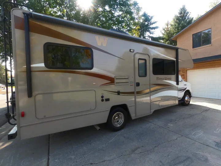 This rv rental, a beige Winnebago Minnie Winnie motorhome, is parked on a sunny concrete driveway next to a house.. Winnebago Minnie Winnie 2014