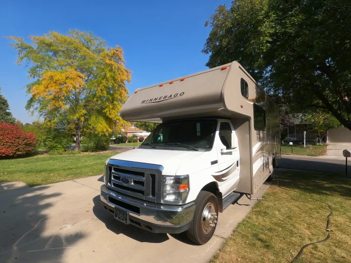 A white and tan Winnebago RV rental sits on a driveway under a clear sky, surrounded by colorful autumn trees.. Winnebago Minnie Winnie 2014