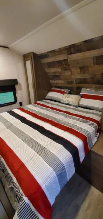Cozy RV rental bedroom with a comfy striped bed, wood accent wall, and bright window for serene views.. Dutchmen Aspen Trail 26BH 2021
