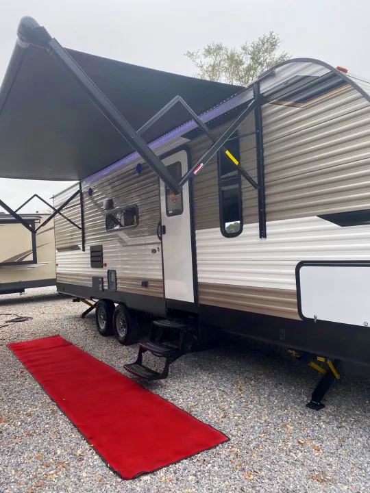 Spacious RV rental travel trailer with extended awning and LED lights, set on a gravel lot with a red mat for comfortable camping.. Dutchmen Aspen Trail 26BH 2021