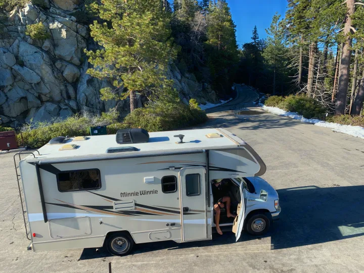 Minnie Winnie RV rental in a mountain setting with a person exiting. Winding road, pine trees, and snow patches are visible.. Winnebago Minnie Winnie 2019